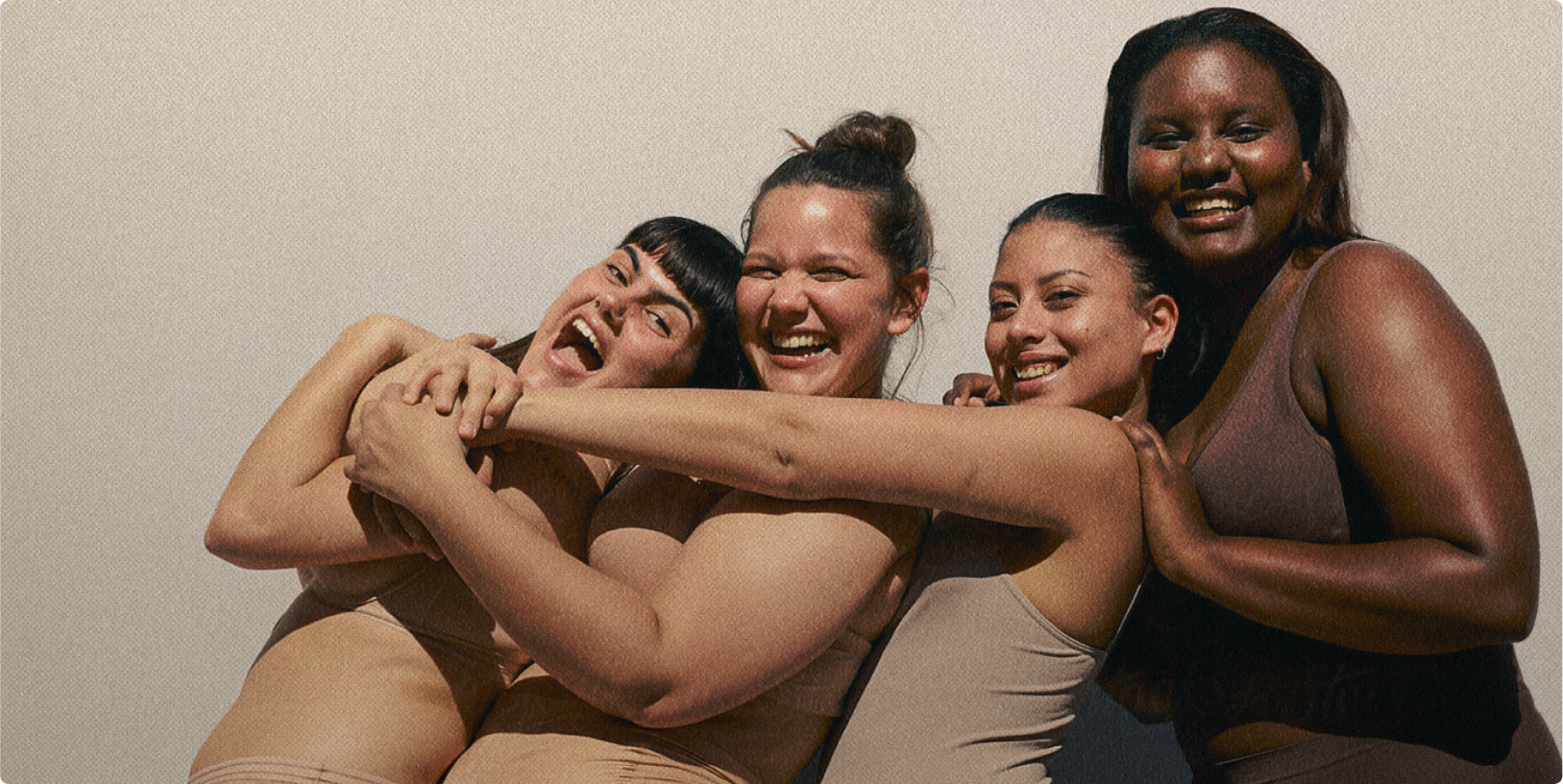 Photographie de 4 femmes s'enlaçant et souriant, de couleur de peau et de morphologie différentes